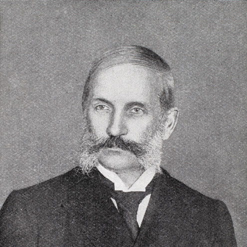 Black and white portrait photo of a young looking John Thomson from 1898. Thomson has a mutton-chop style beard, with his long dark moustache joining up with even longer white sideburns. He is wearing a dark suit, with a white shirt and tie.