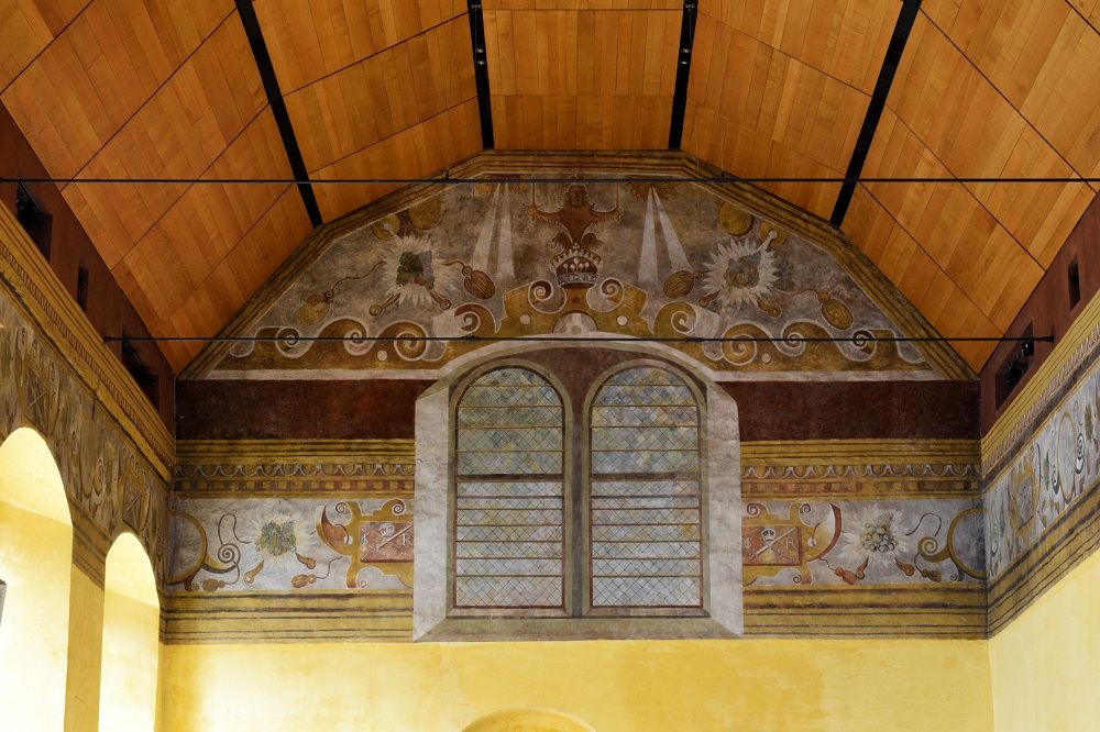 The upper part of a tall room, with large, arched windows. The top of th walls are painted with a mural featuring windows, waves and other designs. The ceiling is arched and clad in wood.