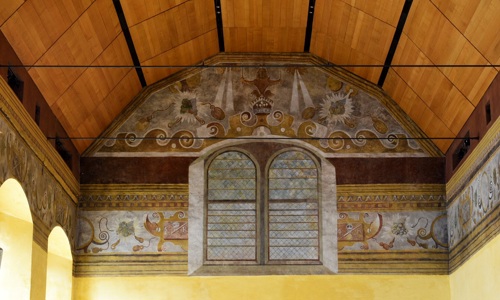 The upper part of a tall room, with large, arched windows. The top of th walls are painted with a mural featuring windows, waves and other designs. The ceiling is arched and clad in wood.