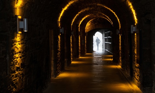A long, arched stone corridor flanked with lights. At the end o f the corridor, an open door leads onto a courtyard, and a person stands in the door.