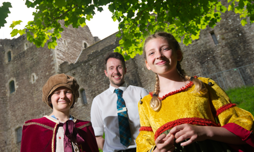 Two Junior tour guide children in medieval period costumes standing on either side of a uniformed member of Historic Scotland staff with a castle in the background.