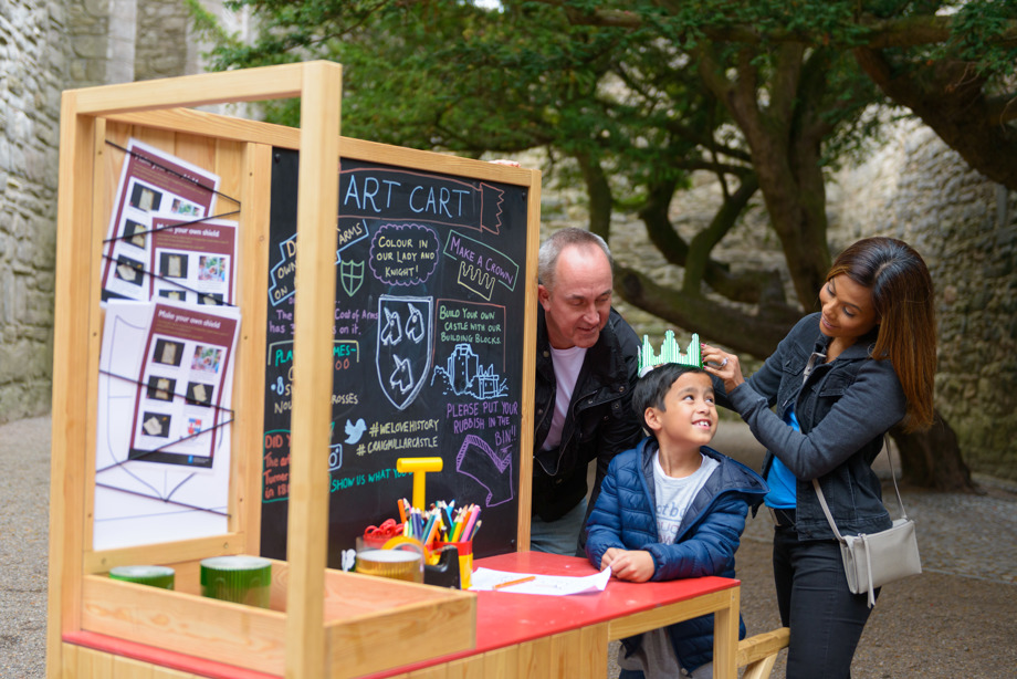 A mum, dad and young boy are doing craft activities at the art cart (a craft station with blackboard, pencils, paper activities).