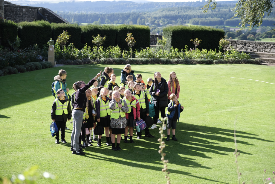 A group of children in hi vis vests are gathered together in a garden whilst an adult is pointing at something.