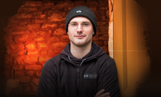 A man wearing an HES fleece and hat, leaning against a brick wall 