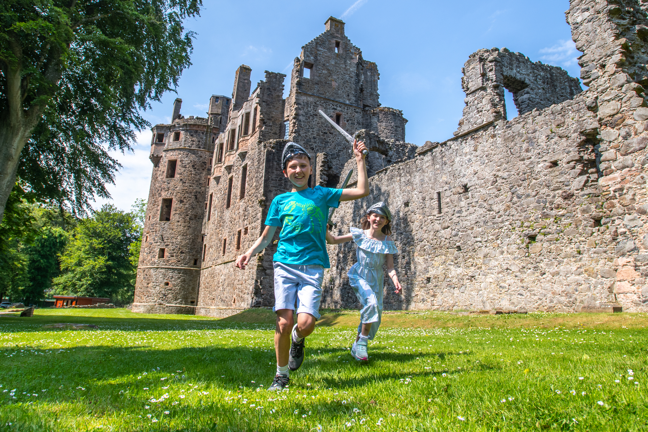Children run with toy swords and helmets in grassy area outside Huntly Castle