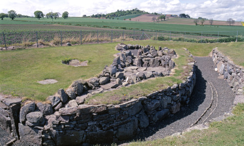 A series of stone walls, partly filled in with soil and covered in grass. Fields in the background.
