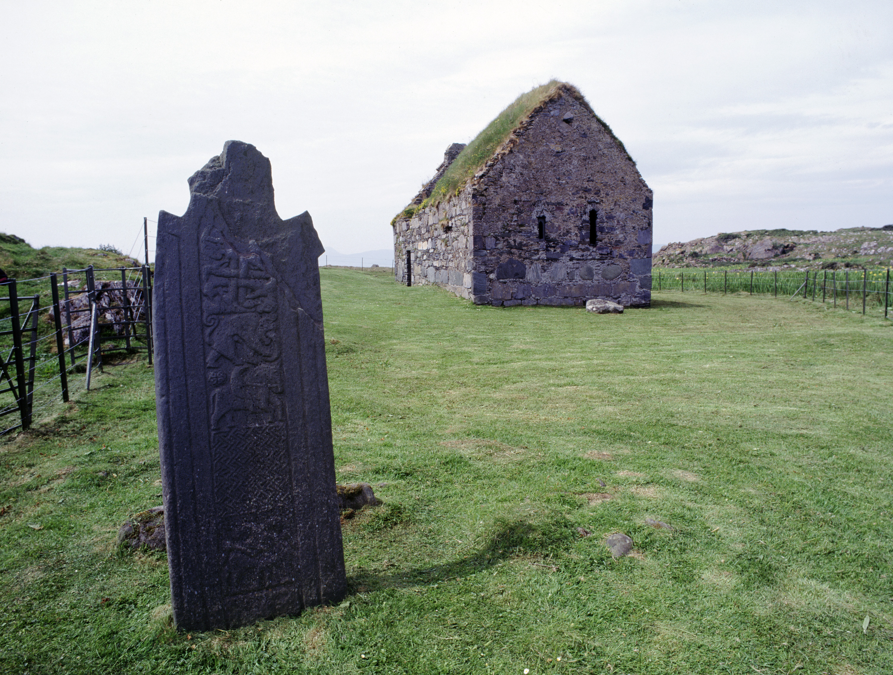 A small stone chapel on grassy ground with a large carved stone in the foreground.