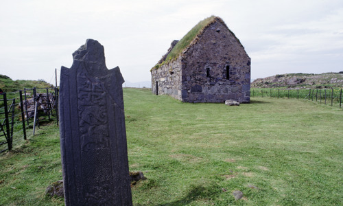 A small stone chapel on grassy ground with a large carved stone in the foreground.