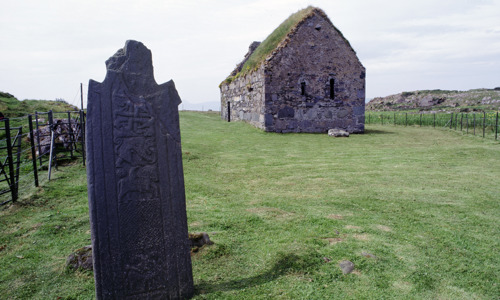 A small stone chapel on grassy ground with a large carved stone in the foreground.