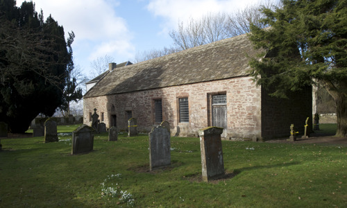 A red stoned cottage surrounded by gravestones and trees.