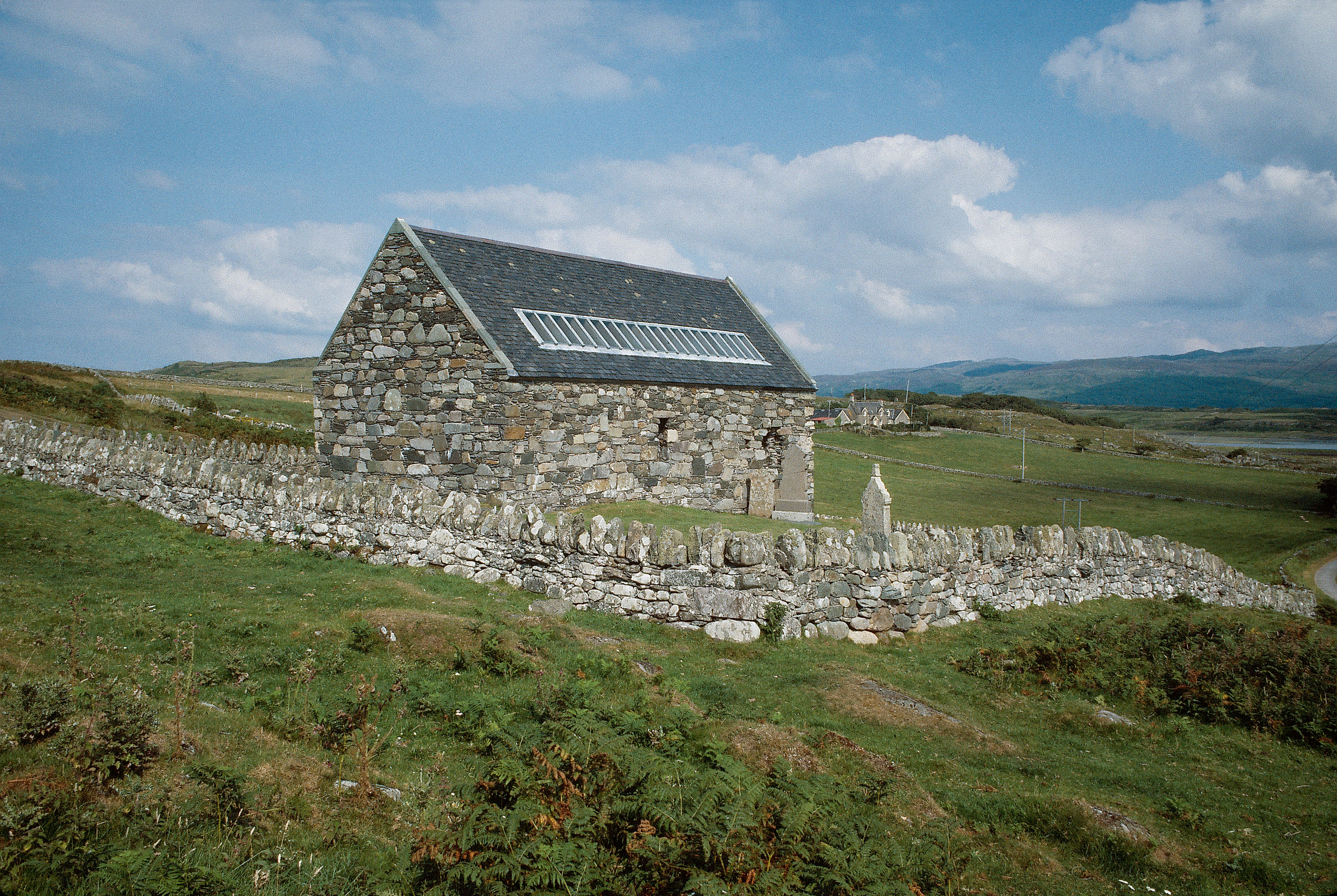 A stone cottage with a silver ladder-like part on the stone roof,  in a rural landscape.