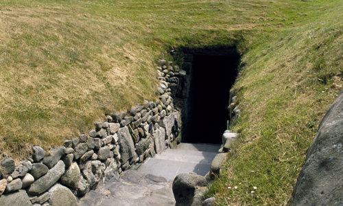 A stone paved path leading into a rectangular opening in a grassy hill.