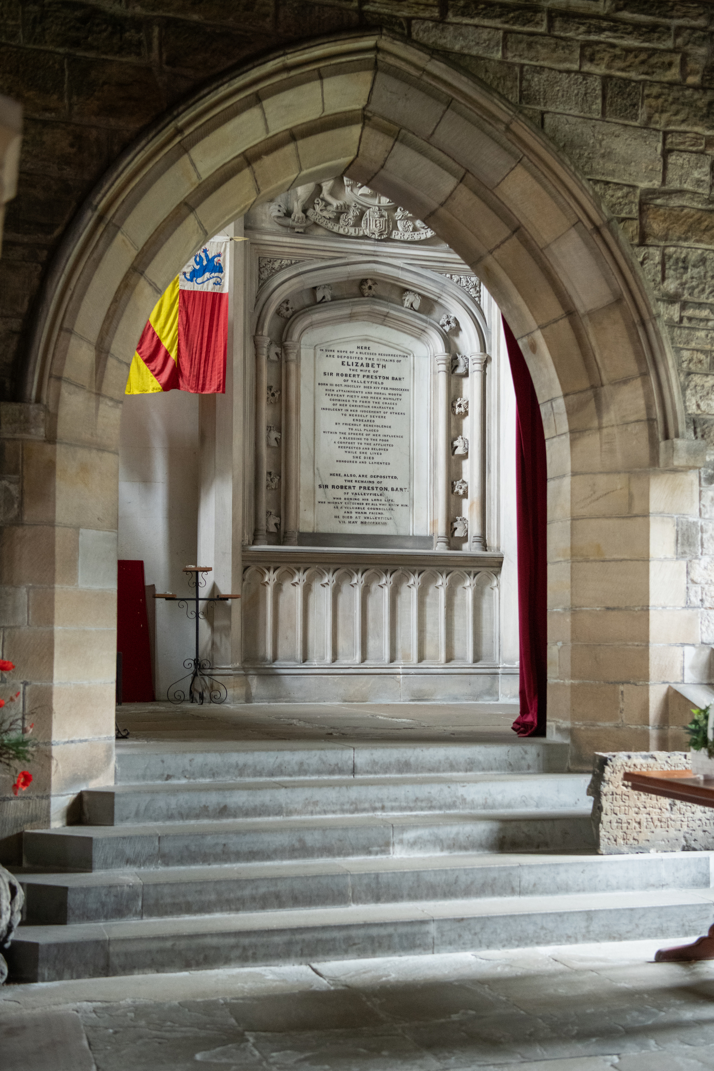A stone archway inside Culross Abbey leading to a memorial plaque carved into an ornate wall, with flags and a small table placed nearby.