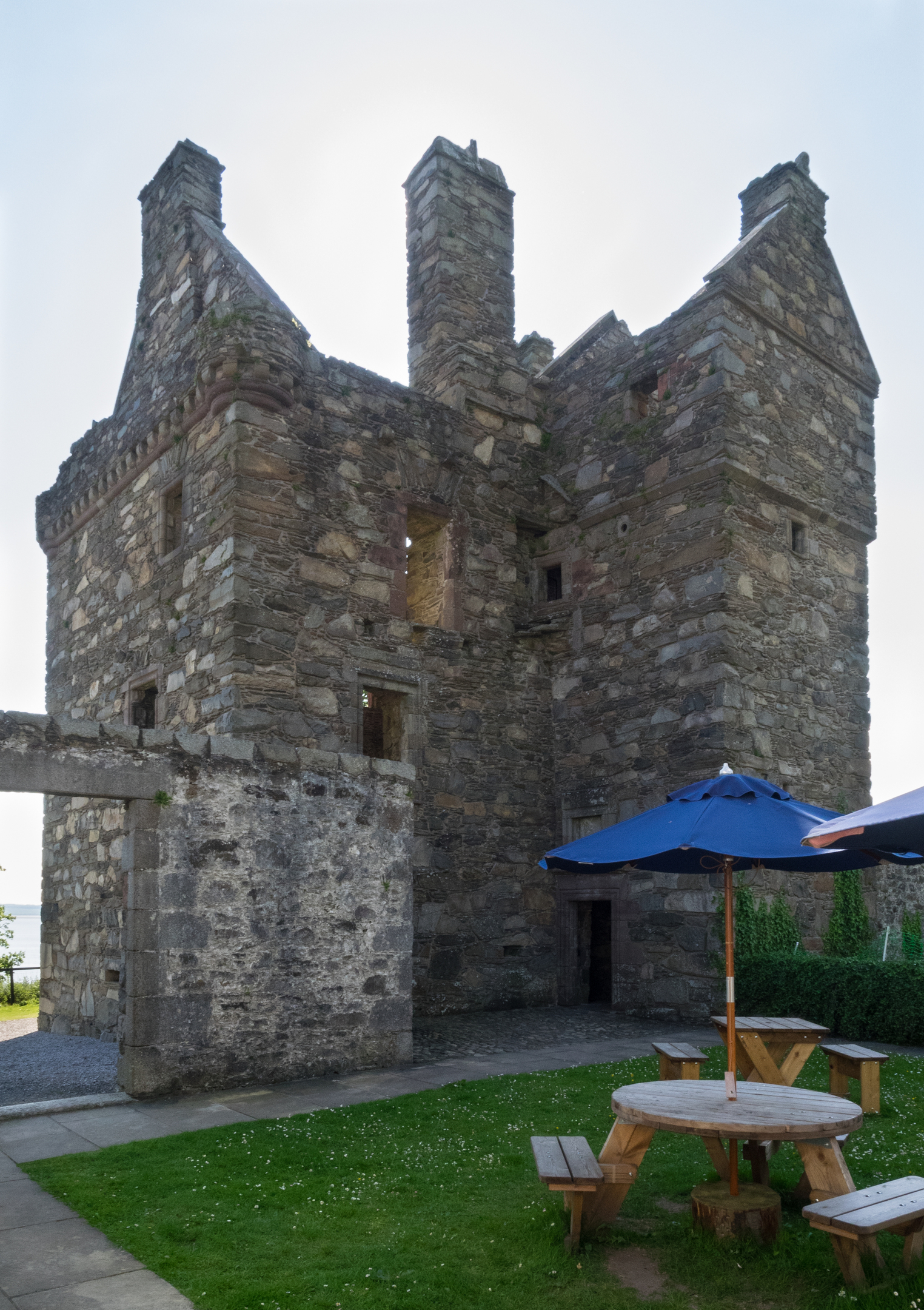 A view of Carsluith Castle showing its tall stone tower house with gabled rooftops and chimneys. In the foreground is a small grassy area with a round wooden picnic table and a blue umbrella.