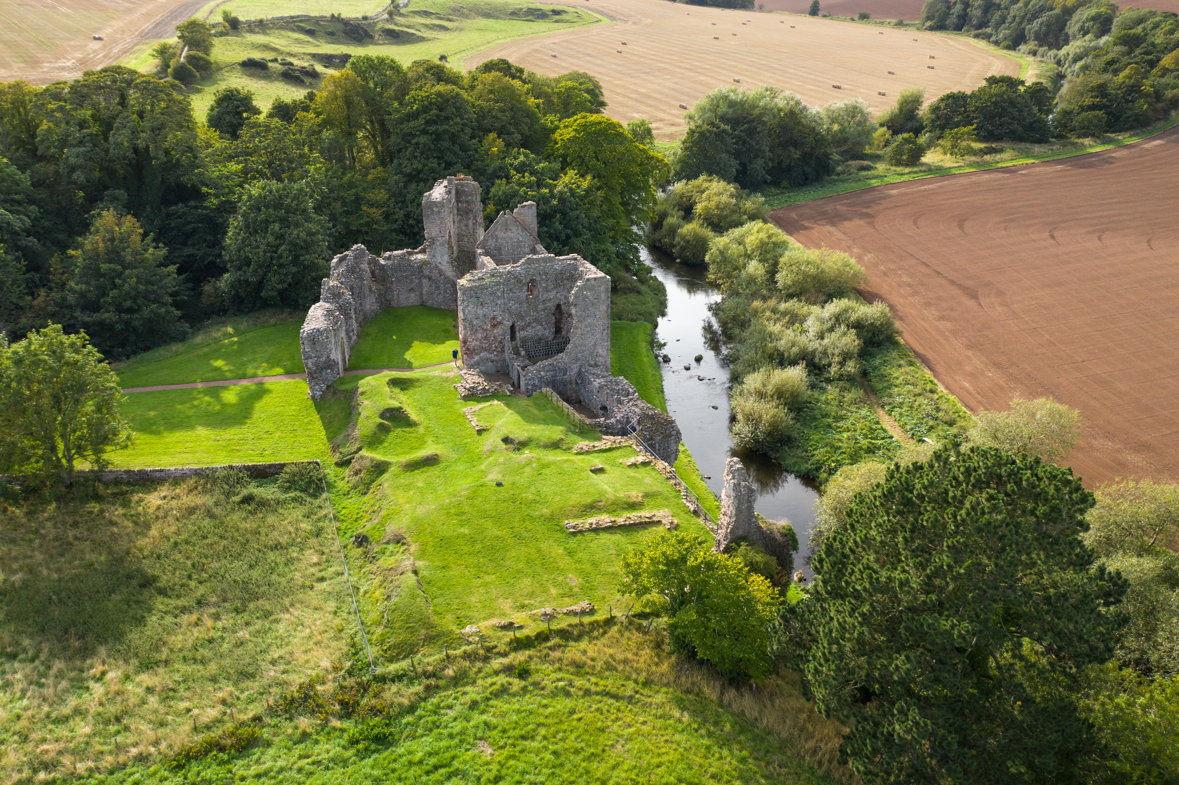 Aerial side view of Hailes Castle ruins perched above a river, with surrounding fields and trees creating a patchwork landscape.