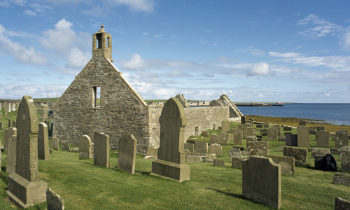 A small roofless chapel with a bell tower surrounded by a graveyard.