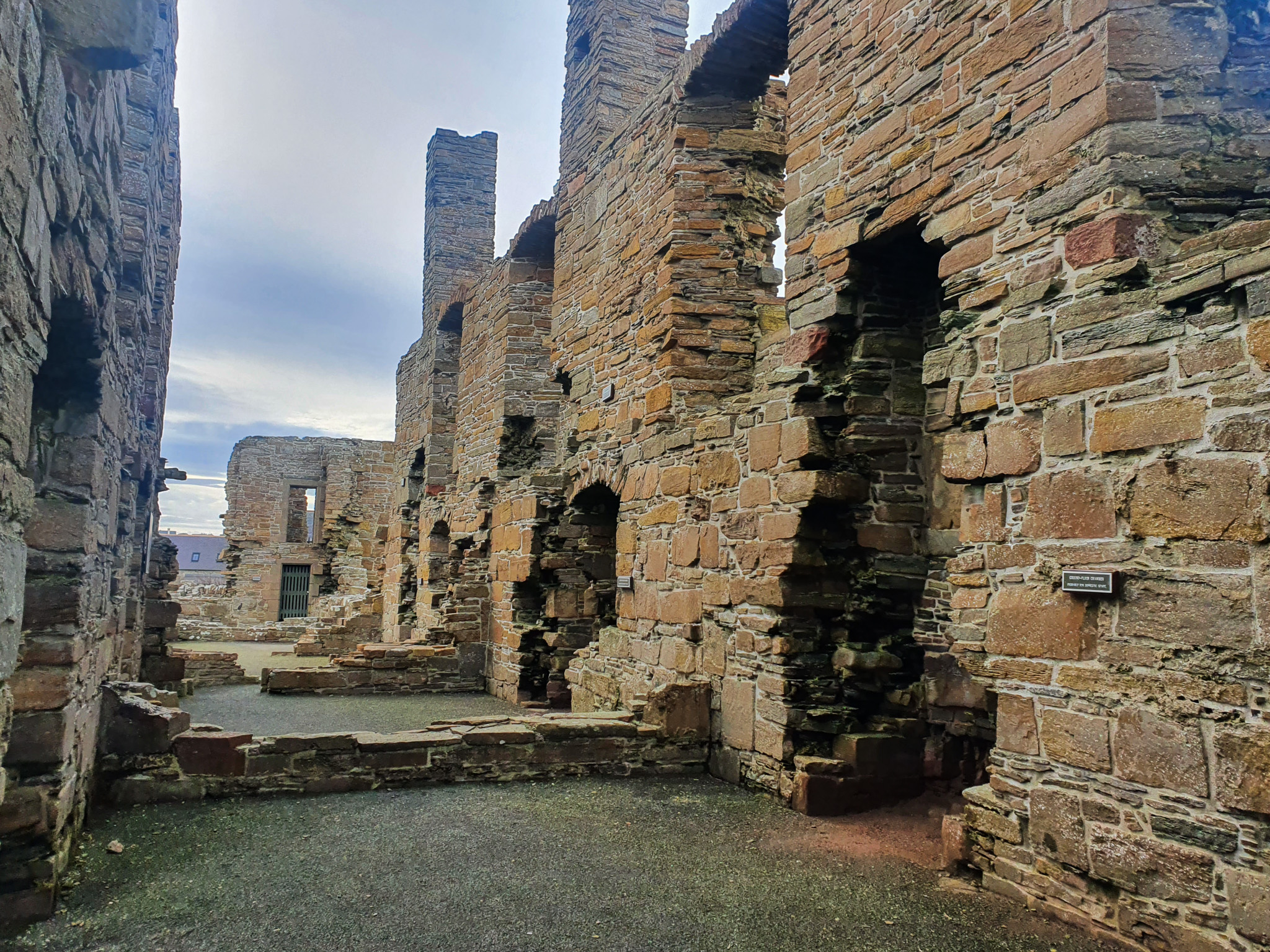 A view inside the ruined Earl’s Palace, showing tall, weathered stone walls lined with empty window and doorway openings. The floor is bare, and the sky is overcast, giving a muted, atmospheric feel to the historic structure.