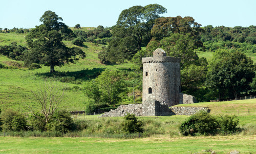 A round stone tower with the foundation of walls on a hilly green landscape