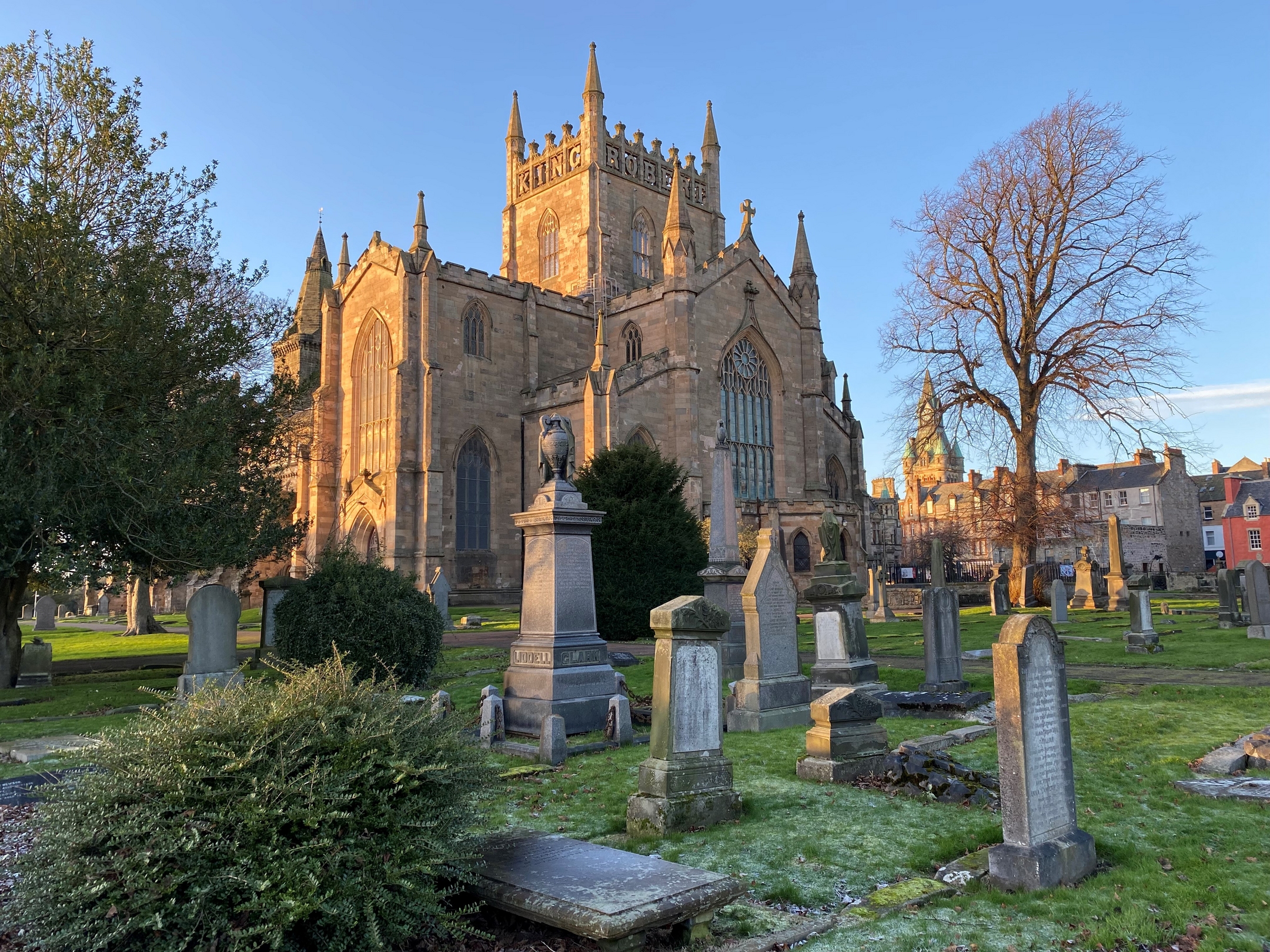 A warm sunrise lights the exterior of a large Gothic-style abbey, surrounded by gravestones and trees, with frost visible on the grass.