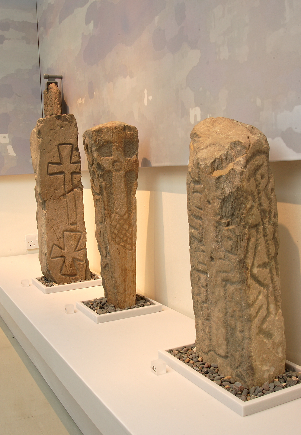 Photograph of three upright carved stones in a museum, each featuring a simple Latin cross or cross‑head on a weathered sandstone surface