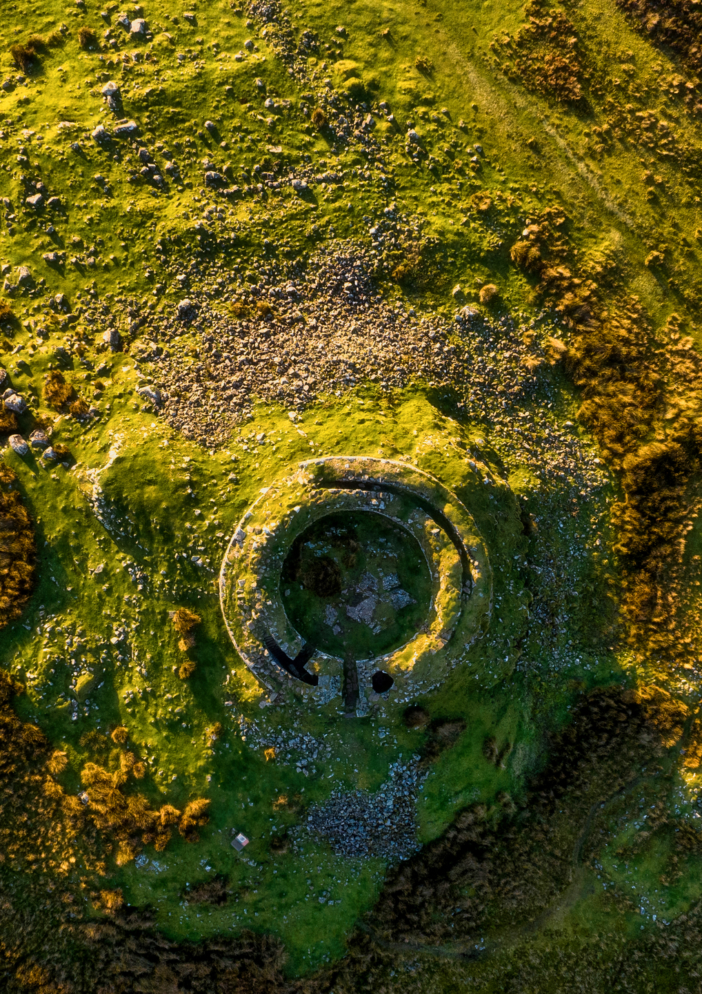 A circular wall made of stone, it is on a mound with grass growing on and around it.