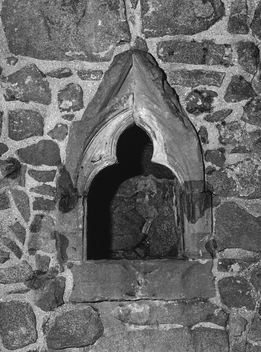 A close‑up black‑and‑white photograph of a small pointed‑arched opening set into a rough stone wall. The arch is formed from carved stone blocks that taper to a central point, with the inner cavity opening into darkness. Weathering marks and surface wear are visible on the carved stones.