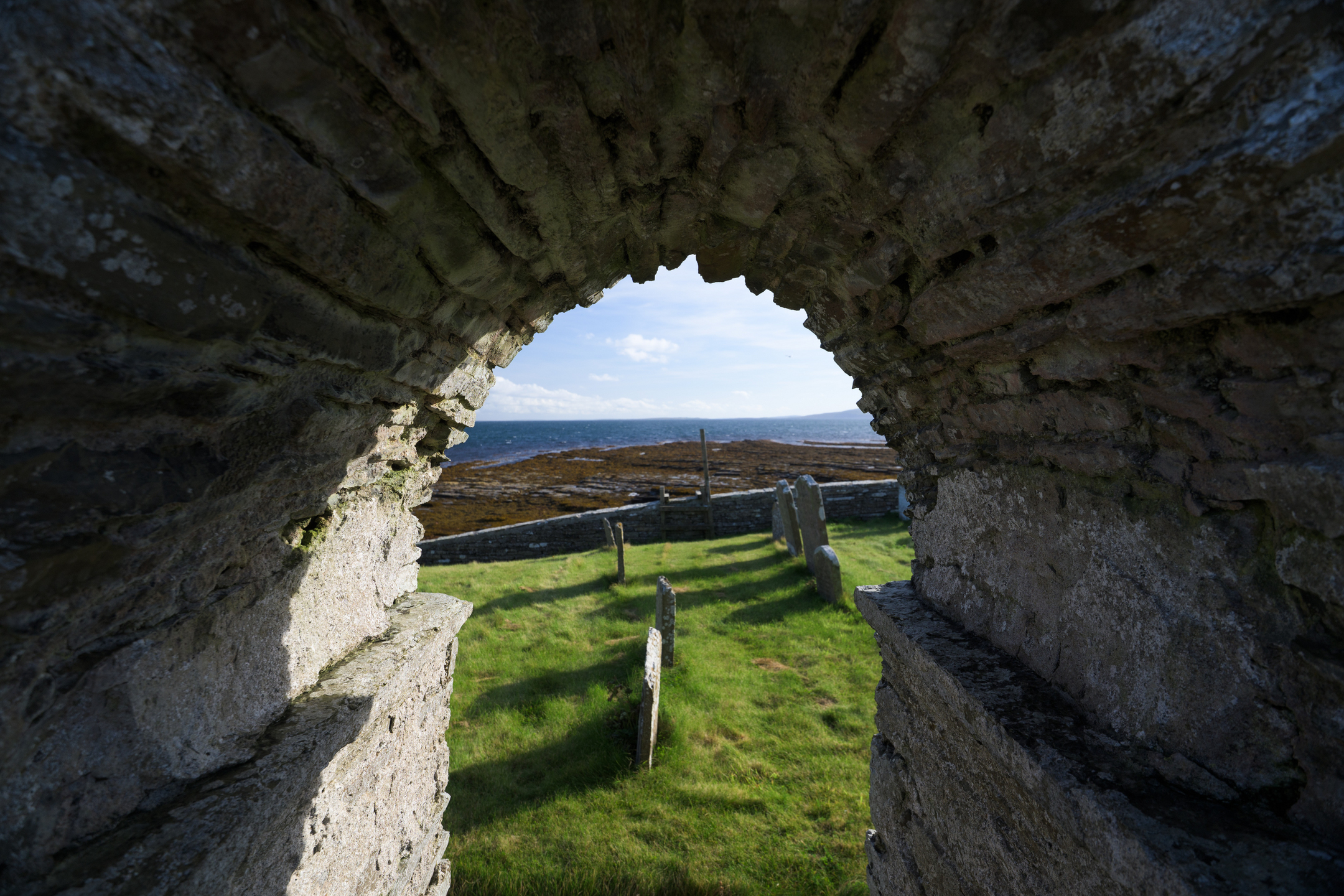 View framed through a stone archway in the church ruins, looking out toward gravestones, green grass, and the distant shoreline.