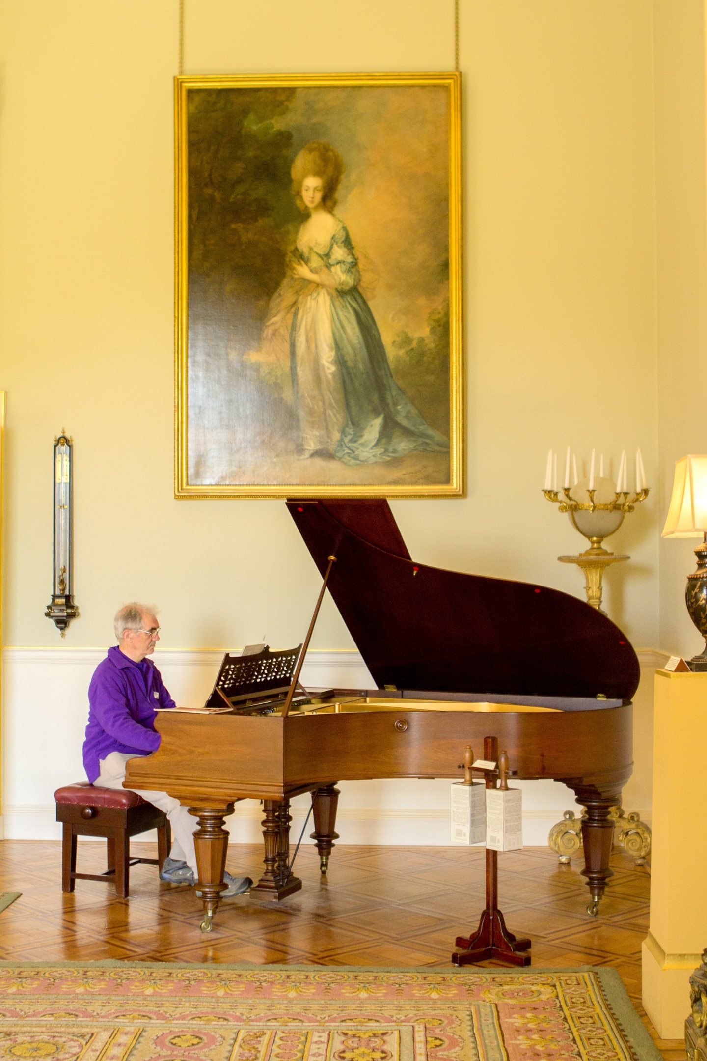 Person seated at a grand piano playing in an elegant room, with a large portrait painting hanging on the wall behind.