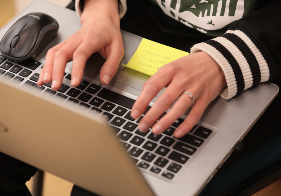 Overhead view of someone using a laptop, with their hands over the keyboard and Post-it note stuck to the mousepad.