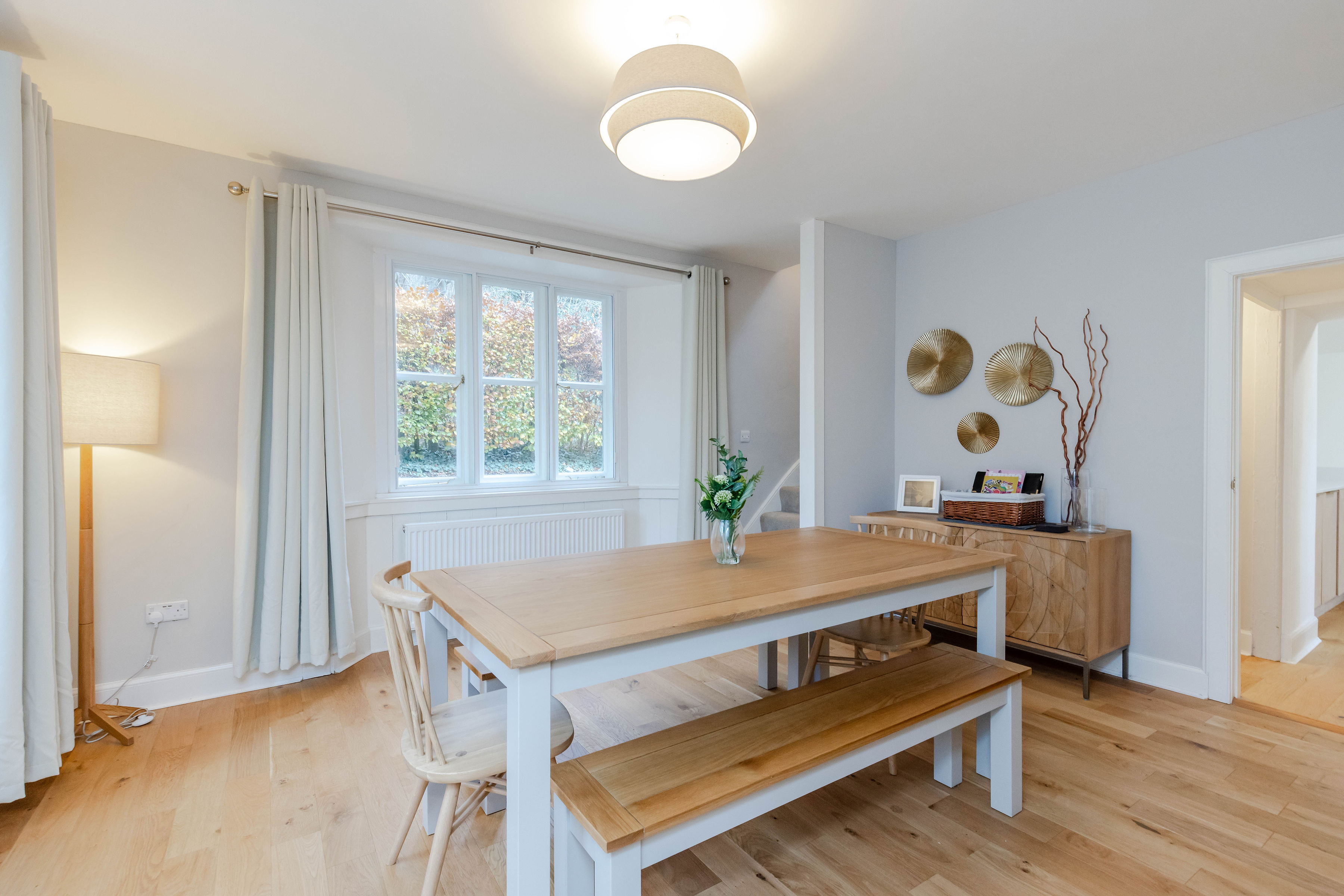 A long, wooden topped dining table flanked with two chairs and two long benches, in a nice, tidy room with a view of a green hedge out the window