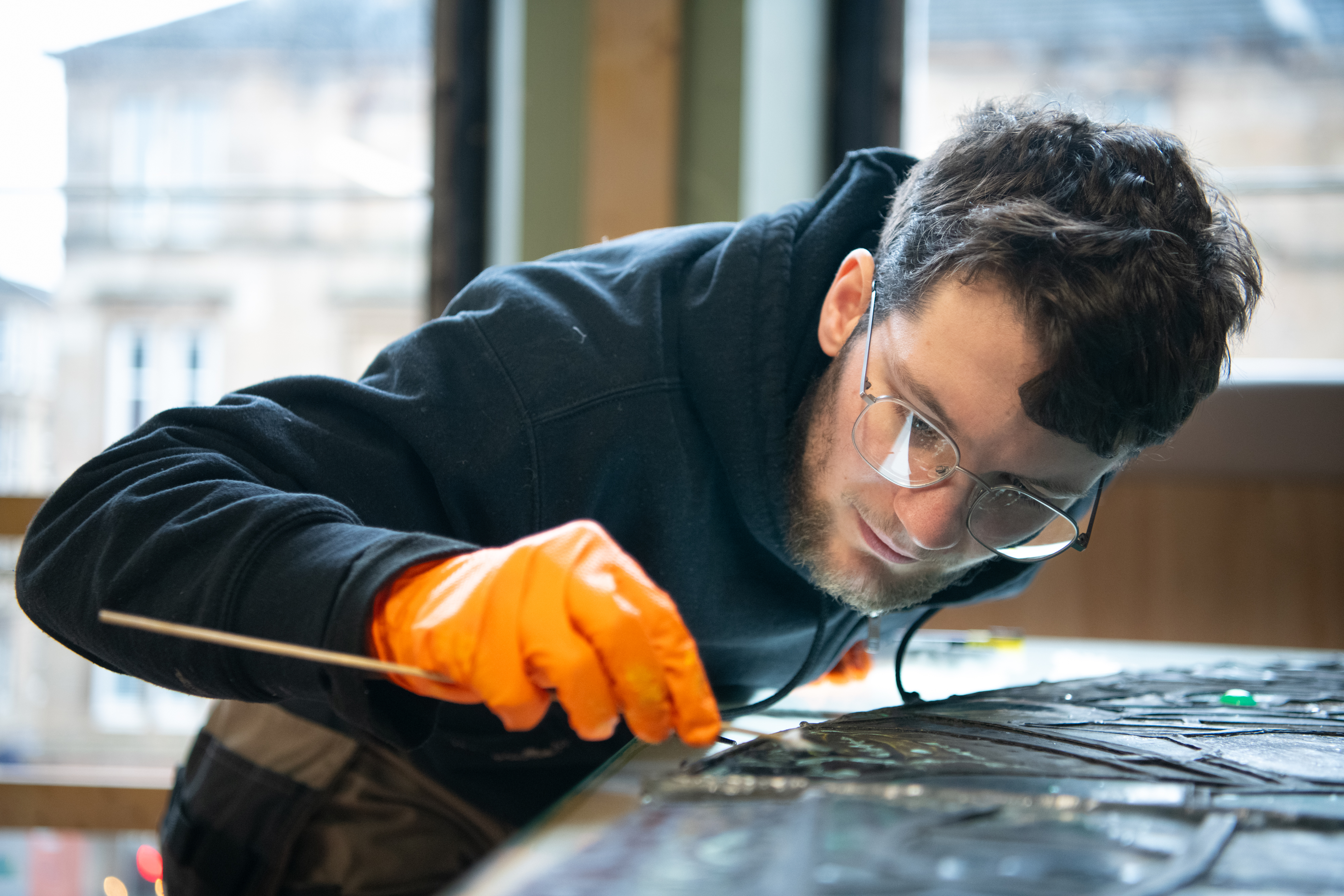 A man wearing an orange glove uses a fine tool to clean a stained glass window on a workbench