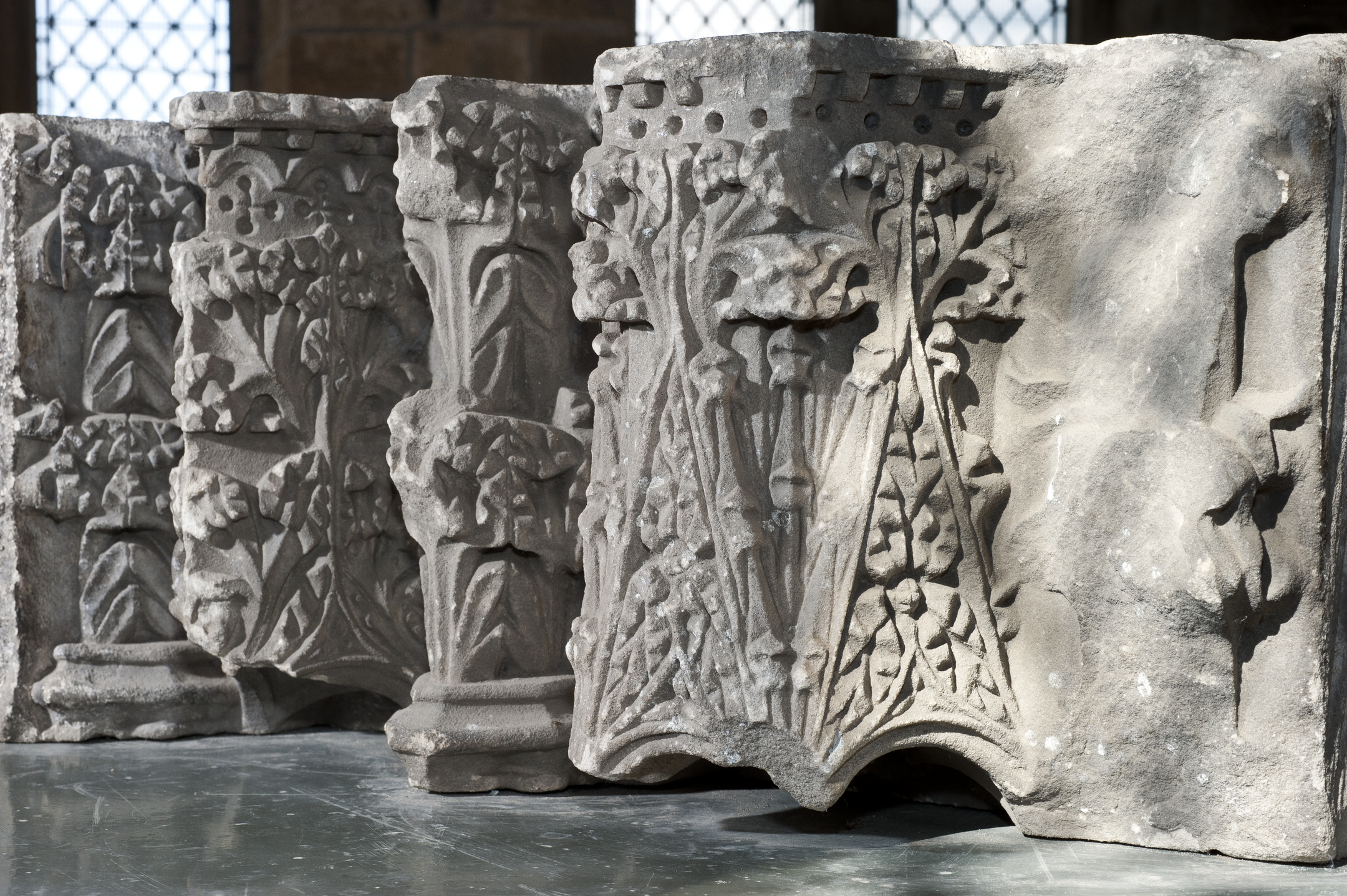 A row of medieval carved stone fragments displayed indoors, each decorated with intricate floral and foliage patterns. The carvings include stylised leaves, stems, and ornamental motifs, illuminated by natural light from nearby windows.