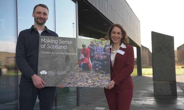 A man and woman pose in front of a modern glass and metal building whilst holding a large board showing a photograph of children exploring a historic site. The board is printed with the words Making Sense of Scotland Outreach and Learning Framework. The sun is shining.