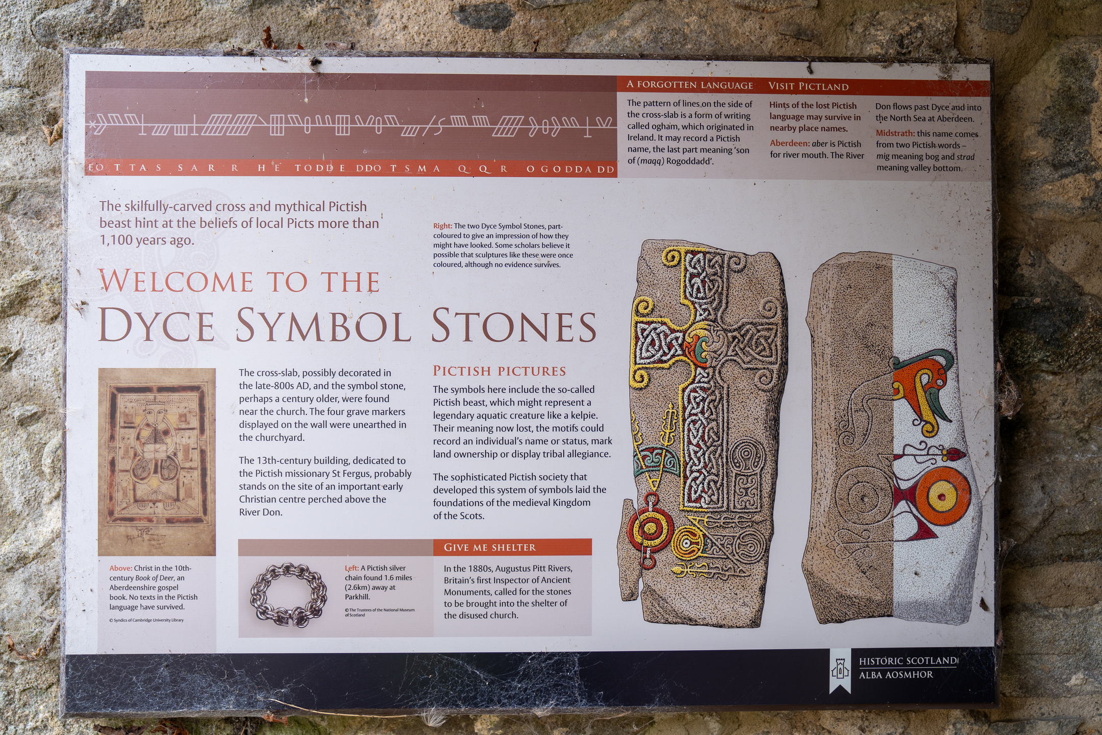 Interpretive sign titled ‘Welcome to the Dyce Symbol Stones’ mounted on a stone wall. The panel includes text explaining the significance of Pictish carvings, illustrations of two symbol stones with intricate designs including a cross and mythical beasts, and a section about the Pictish language and history. Decorative motifs and color highlights emphasize key details.