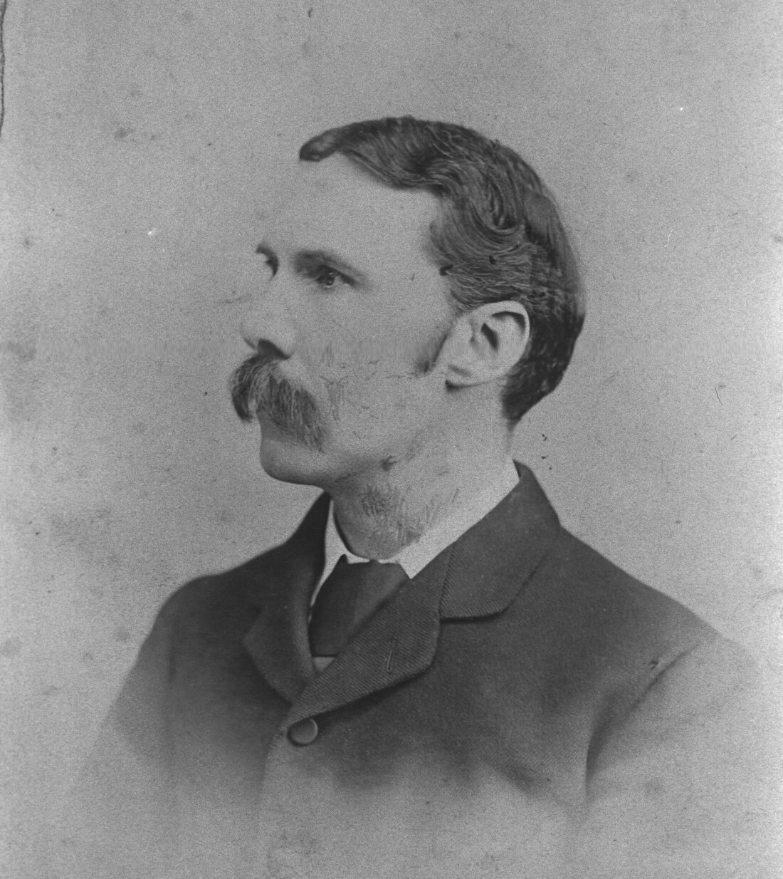 Black and white portrait photo of a person looking to the left, wearing a dark suit and tie. They have a large handlebar moustache.