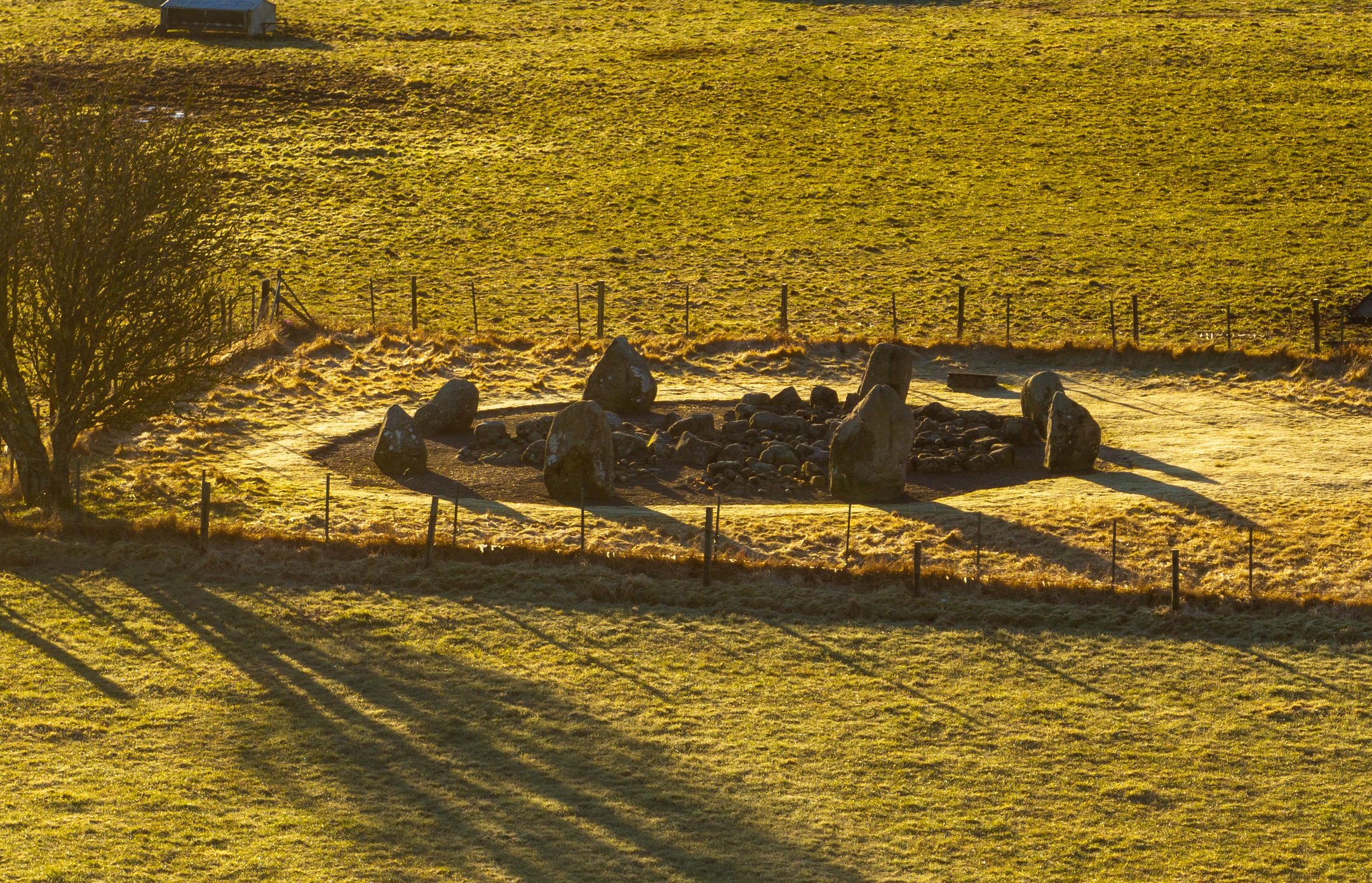 Aerial view of Cullerlie Stone Circle in golden sunrise light, showing a ring of large standing stones surrounding smaller cairn mounds within a fenced grassy field.
