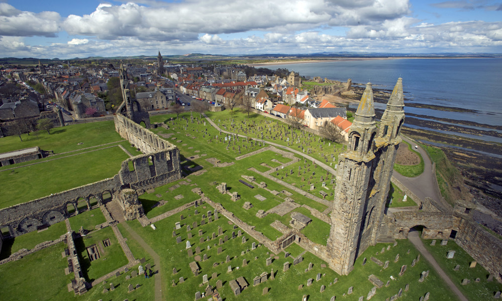 An aerial photograph looking down at the ruin of a cathedral set within a graveyard beside a small town and a bay.