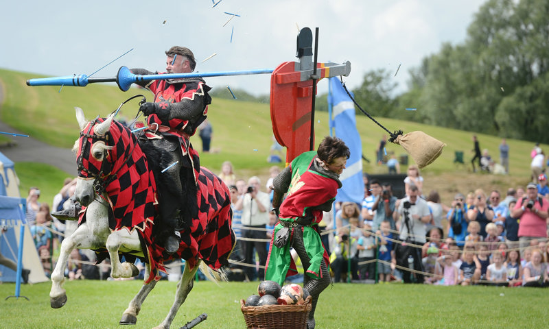 Action shot of a jouster on horseback assaulting a wooden cutout of a knight, with splinters flying through the air