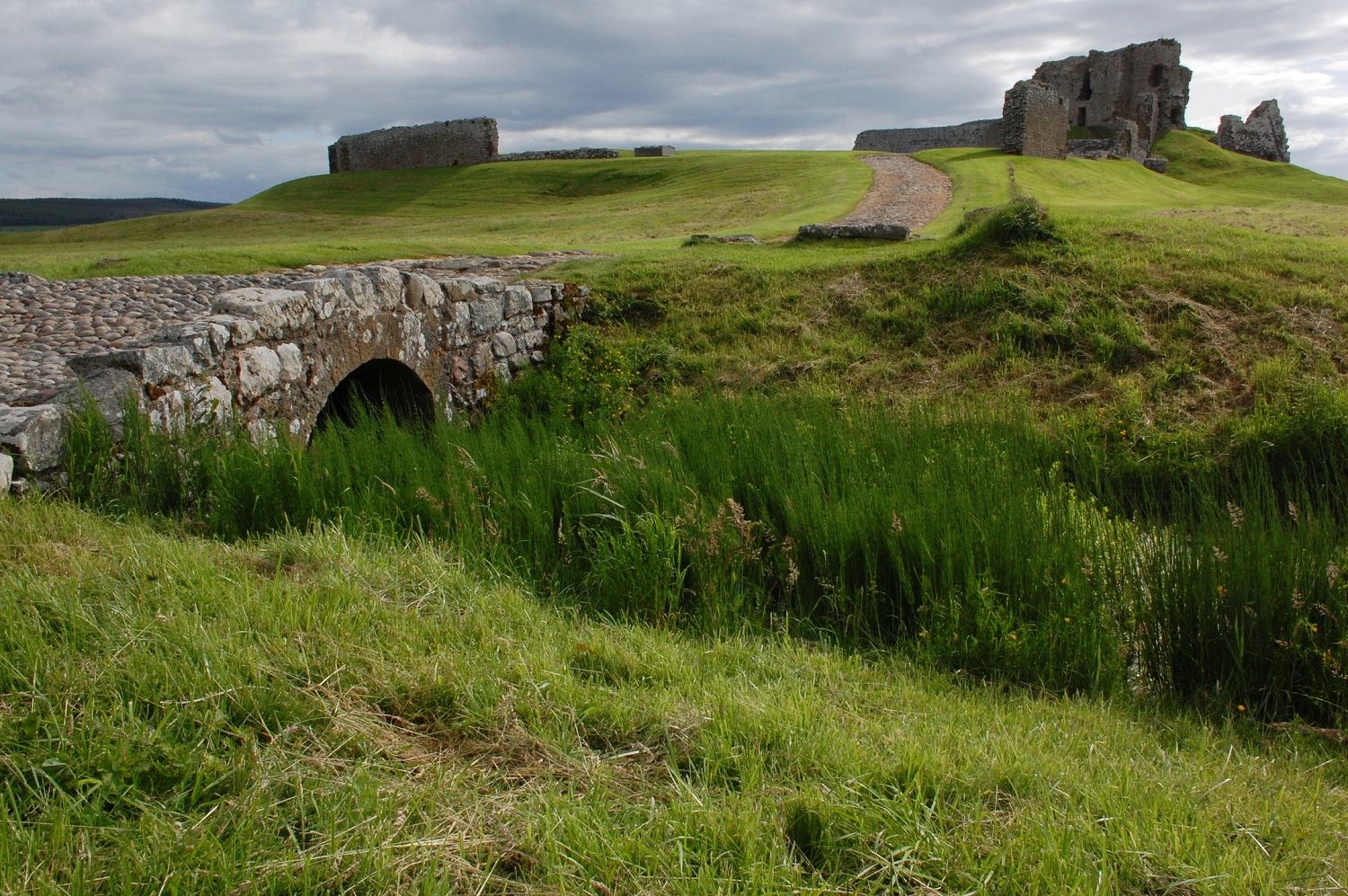 Exterior view of Duffus Castle ruins on a grassy hill beneath a wide sky. A small stone foot bridge is in the foreground.
