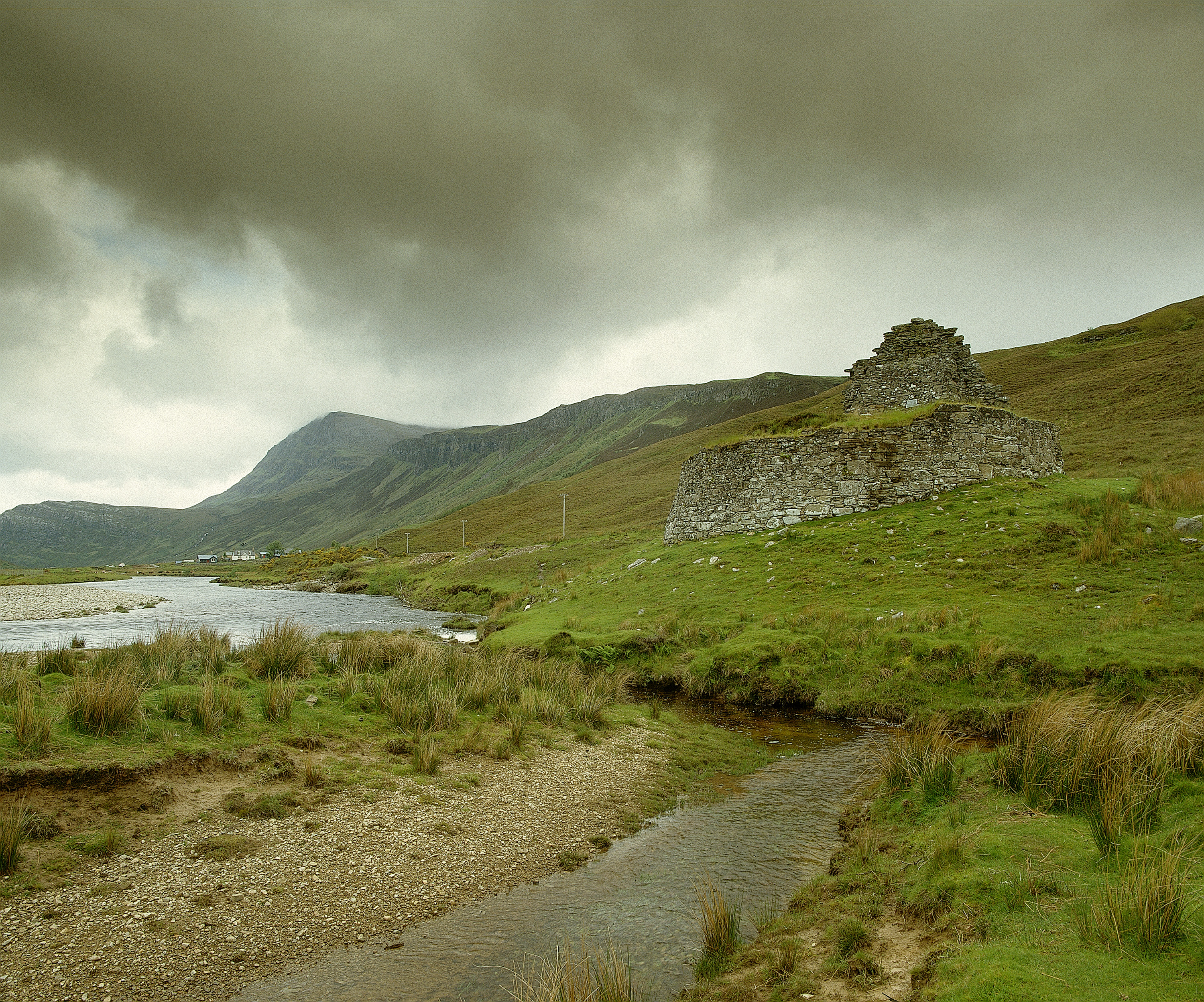 Rear view of the ruined stone broch at Dun Dornaigil, standing on a grassy hillside beside a winding river. The circular stone structure is partially collapsed, with uneven upper walls. In the background are rolling hills and mountains under heavy, overcast clouds that cast a muted light across the landscape.