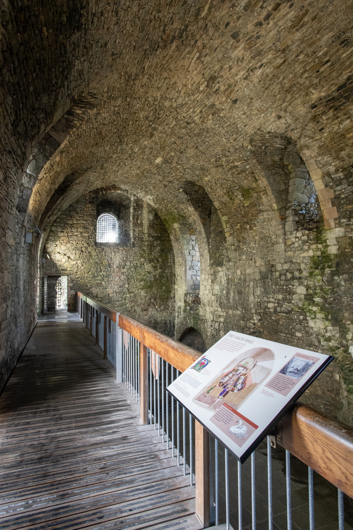 An interior walkway inside the castle, showing a long arched stone corridor with narrow windows and an information panel along a wooden platform with railings.