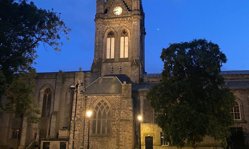 View of a church by night with lights on and gravestones and trees in the foreground