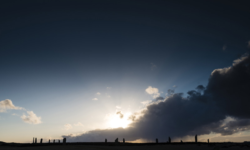A distant photo of the sun going down behind the Ring of Brodgar.