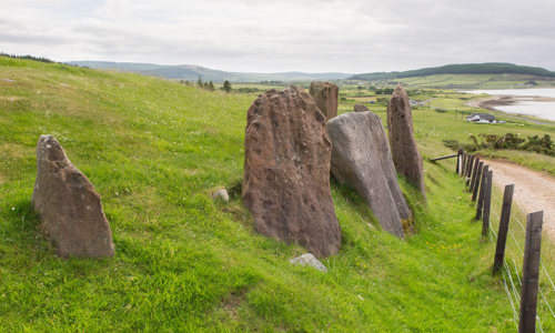 Four medium sized standing stones are set in a row next to a small road. The sea can be seen in the distance.
