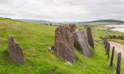 Four medium sized standing stones are set in a row next to a small road. The sea can be seen in the distance.