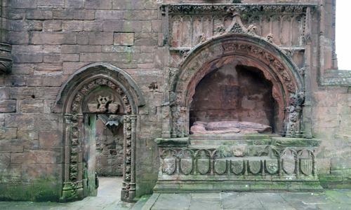 A stone wall with a doorway on the left and a lavishly decorated tomb on the right