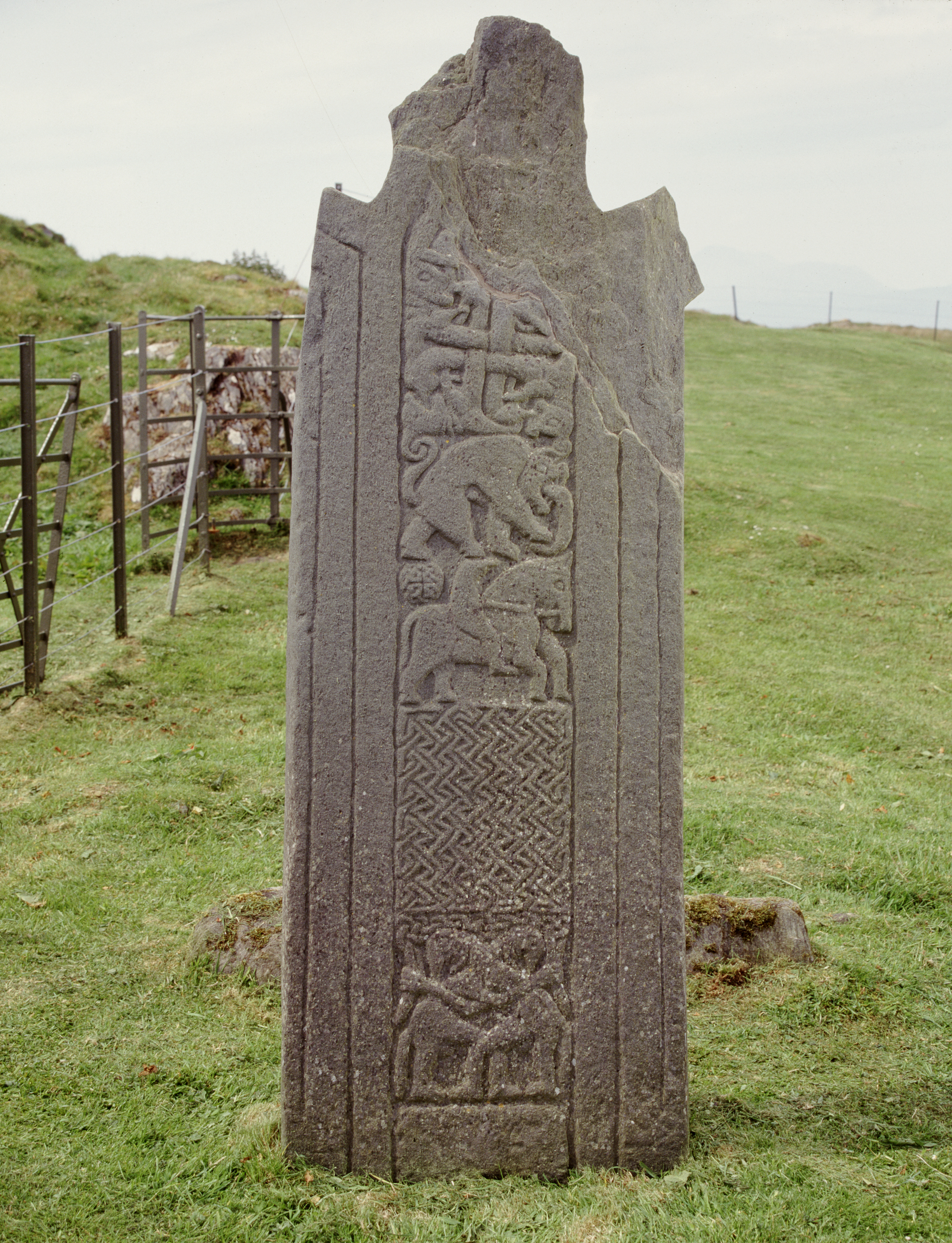 Weathered stone cross shaft with intricate carvings of animals and interlaced patterns, standing on grass near a metal fence.