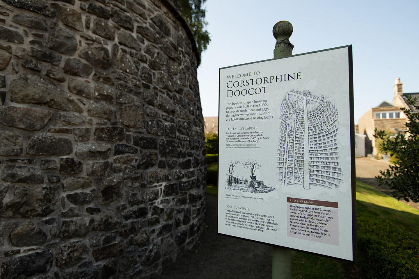 Sign next to a historic stone wall which reads "welcome to Corstorphine Doocot" and has an illustration of what it looks like inside, a tall building with lots of stone nesting boxes for doves or pigeons