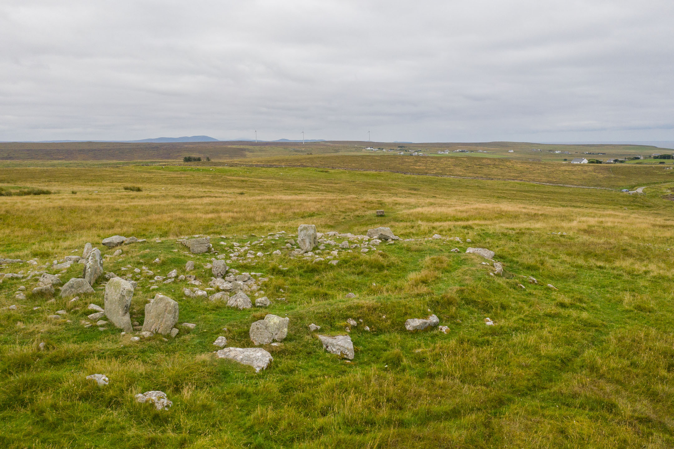 Wide view of cairn and stone circle remains on grassy moorland with distant hills.