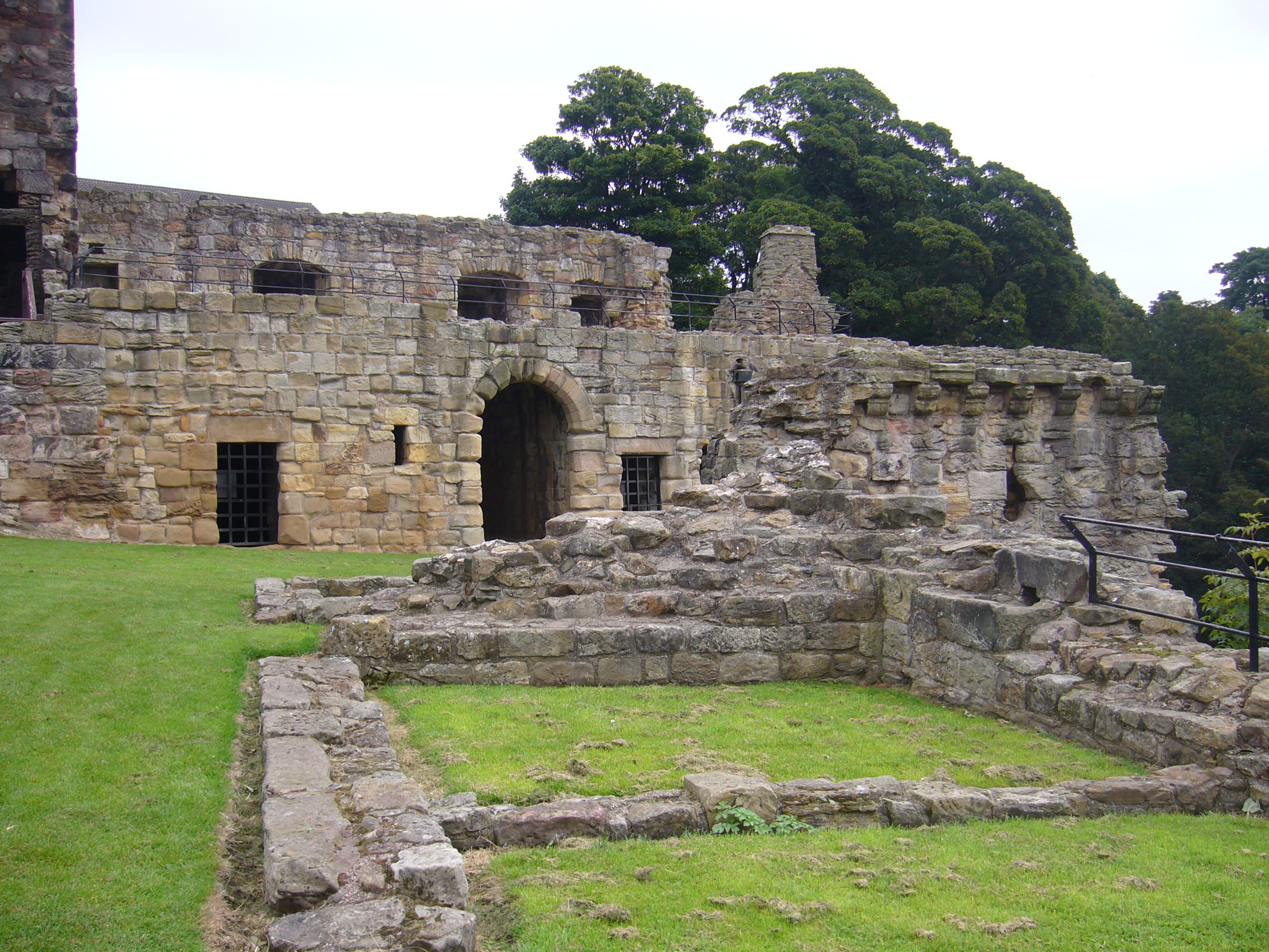 A ground-level view of the interior ruins of Ravenscraig Castle showing thick stone walls, a large arched doorway, and low, crumbling foundations marking former rooms. The stonework is worn and irregular, with barred windows set into the walls. Green grass fills the open courtyard, and tall trees stand behind the ruins.